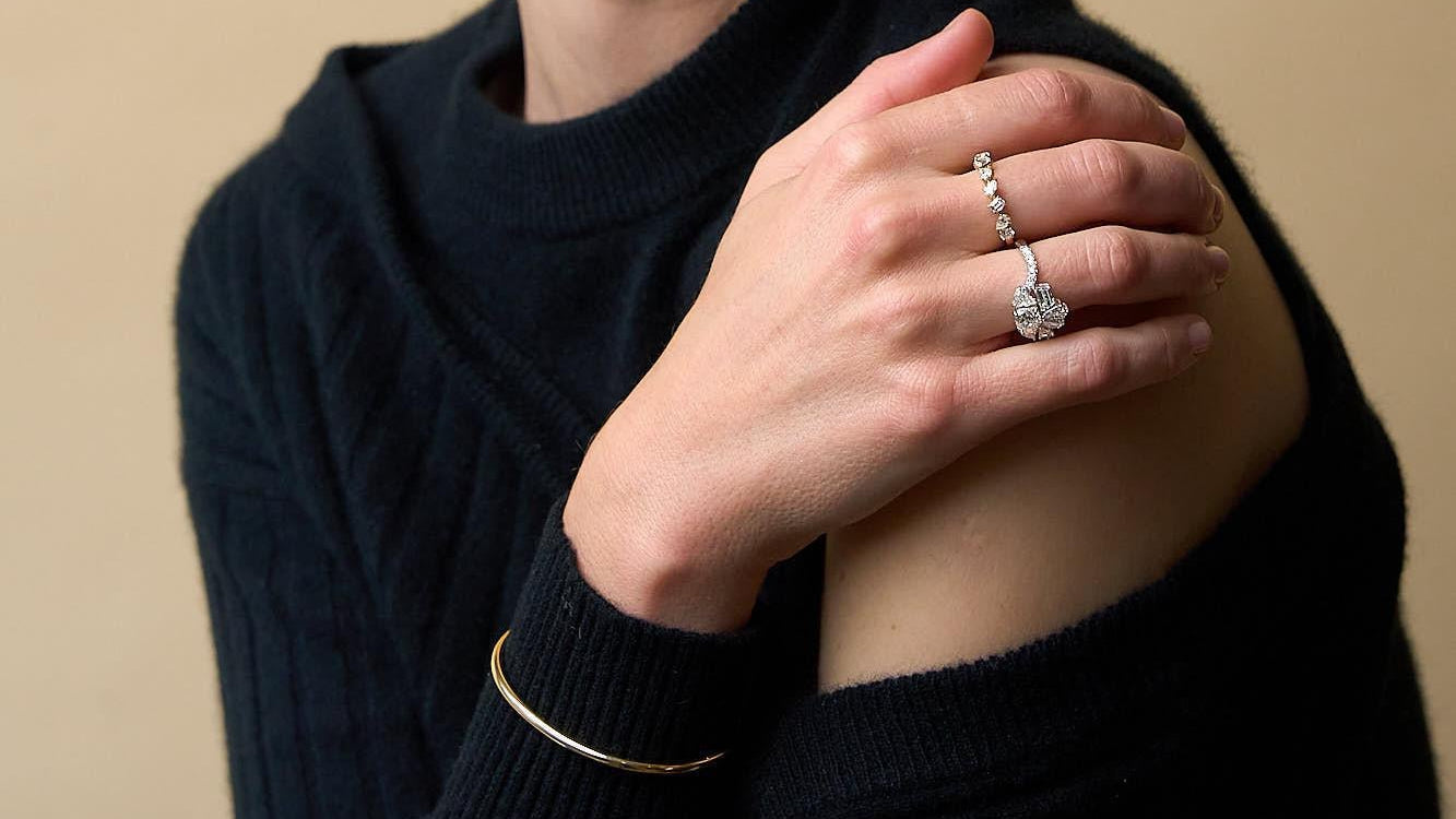 Woman wearing a dark sweater, diamond rings and a gold bracelet against a beige background